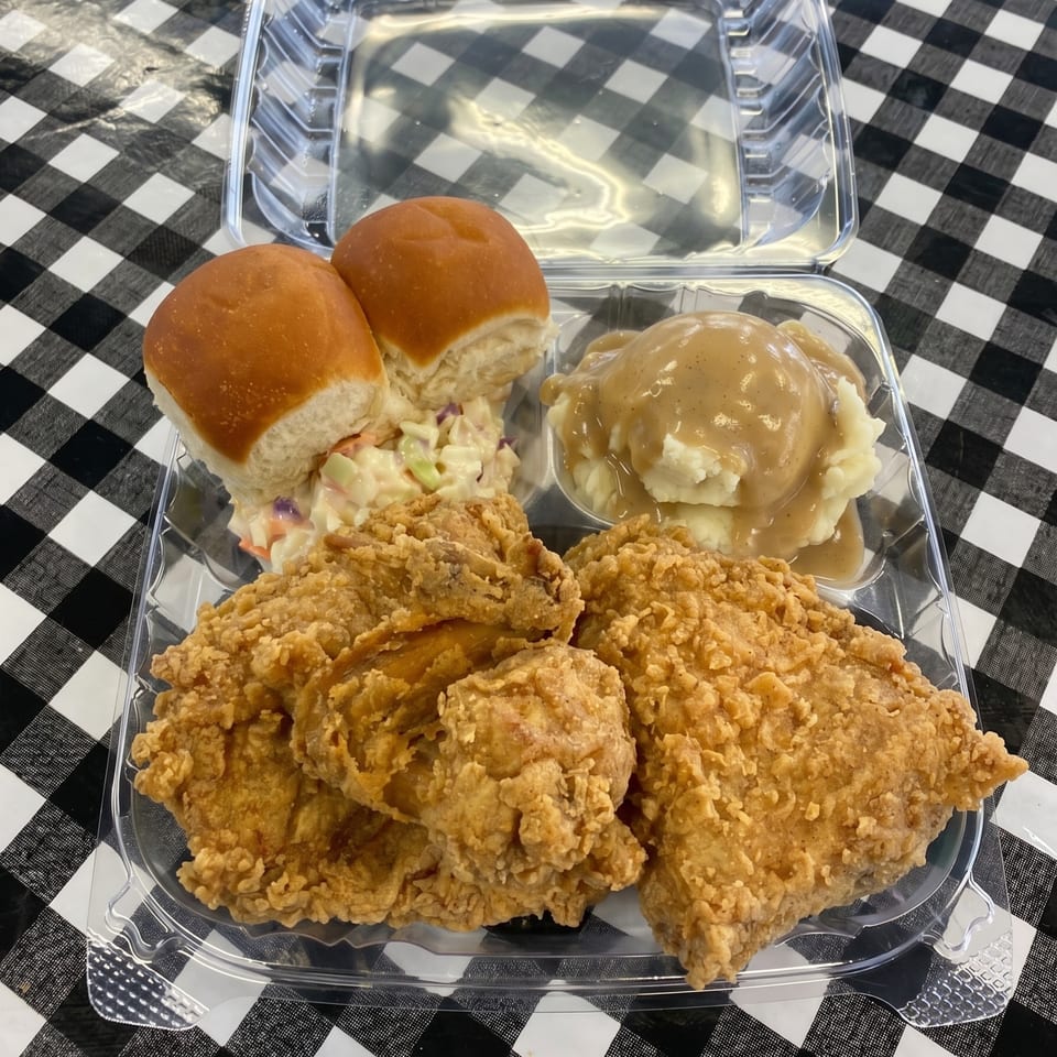 A fried chicken meal with mashed potatoes, gravy, coleslaw, and dinner rolls in a plastic container.