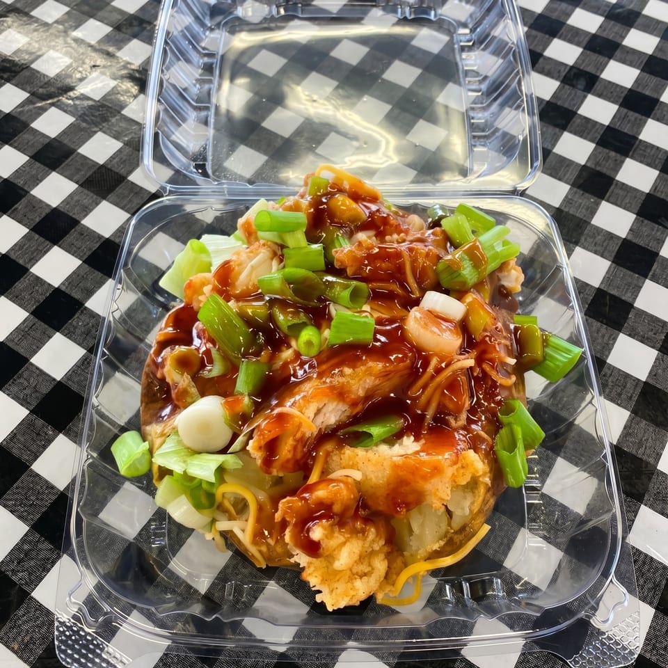 Loaded baked potato with fried chicken, sauce, cheese, and green onions in a plastic container.
